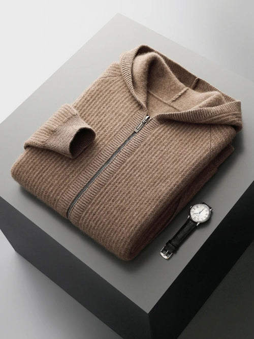 A folded taupe-brown ribbed knit zip-up sweater with a hood, displayed on a dark gray pedestal alongside a black leather strap watch.