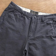 Dark gray chino pants laid flat on a light wood surface, showing the waistband, button closure, belt loops, and front pockets.