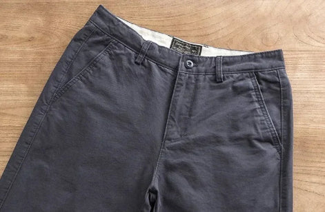 Dark gray chino pants laid flat on a light wood surface, showing the waistband, button closure, belt loops, and front pockets.