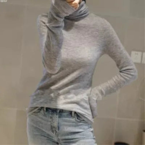 A woman wearing a light gray long-sleeve turtleneck sweater and light-wash blue jeans, shown in a mirror selfie style against a tiled wall.
