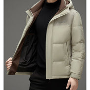 A man wearing a light beige hooded puffer jacket with a brown interior lining over a black turtleneck sweater, shown against a plain gray studio background.