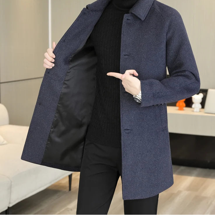 A man wearing a dark navy blue wool-blend coat over a black turtleneck sweater and black pants, standing in a modern indoor room and opening the coat to show the black interior lining.