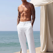 White linen wide-leg trousers worn by a model standing on a beach patio with the ocean and a cream curtain in the background.