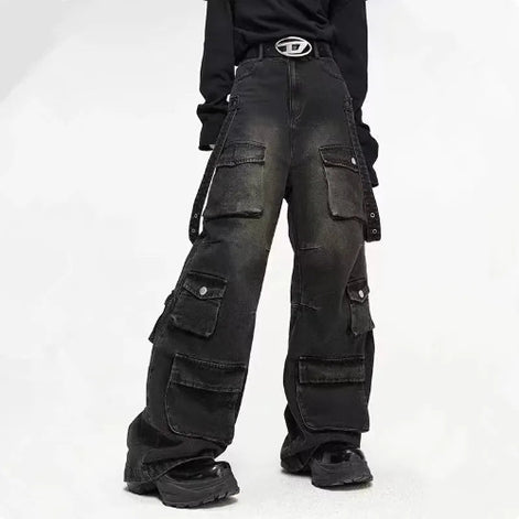 Front view of a model wearing washed black baggy cargo jeans with multiple flap pockets and hanging side straps, paired with black chunky shoes.