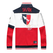 A color-blocked long-sleeve track jacket shown from the back, featuring navy blue, white, and red panels with a shark graphic crest, 'SHARK' text, and sleeve graphics, on a plain white background.