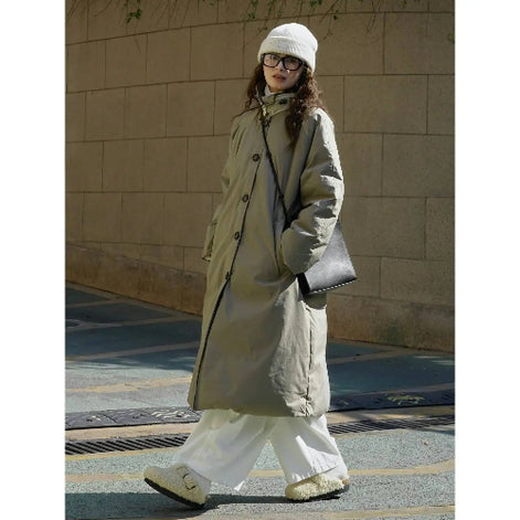 Front three-quarter view of a woman wearing an army green long padded coat with a stand collar and visible front buttons, carrying a black shoulder bag outdoors