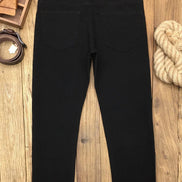 Black slim-fit jeans shown from the back, laid flat on a wooden surface with a belt, leather pieces, and rope as styled props.