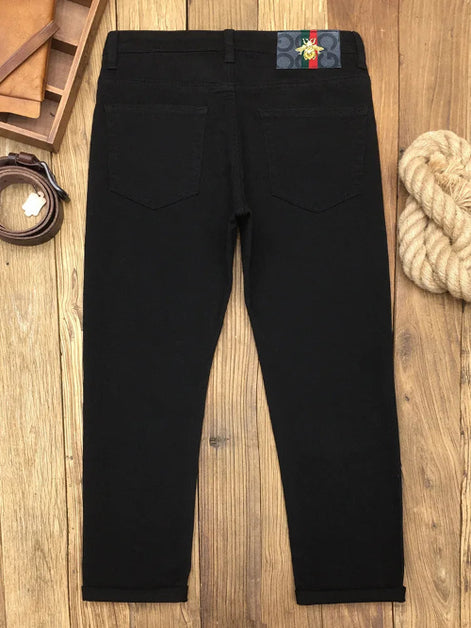 Black slim-fit jeans shown from the back, laid flat on a wooden surface with a belt, leather pieces, and rope as styled props.