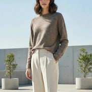 A woman wearing a long-sleeve crewneck knit sweater in a heathered taupe-brown color, paired with black pants, standing indoors against a light curtain background.