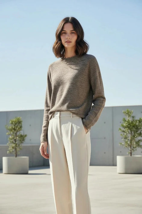 A woman wearing a long-sleeve crewneck knit sweater in a heathered taupe-brown color, paired with black pants, standing indoors against a light curtain background.