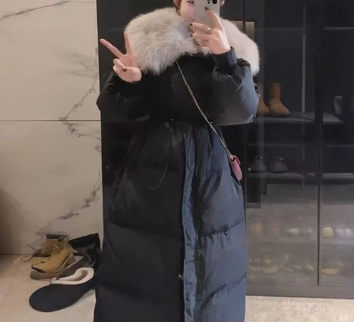 Model taking a mirror selfie in a long black quilted coat with a large cream fur-look collar and cinched waist, wearing a chain-strap crossbody bag