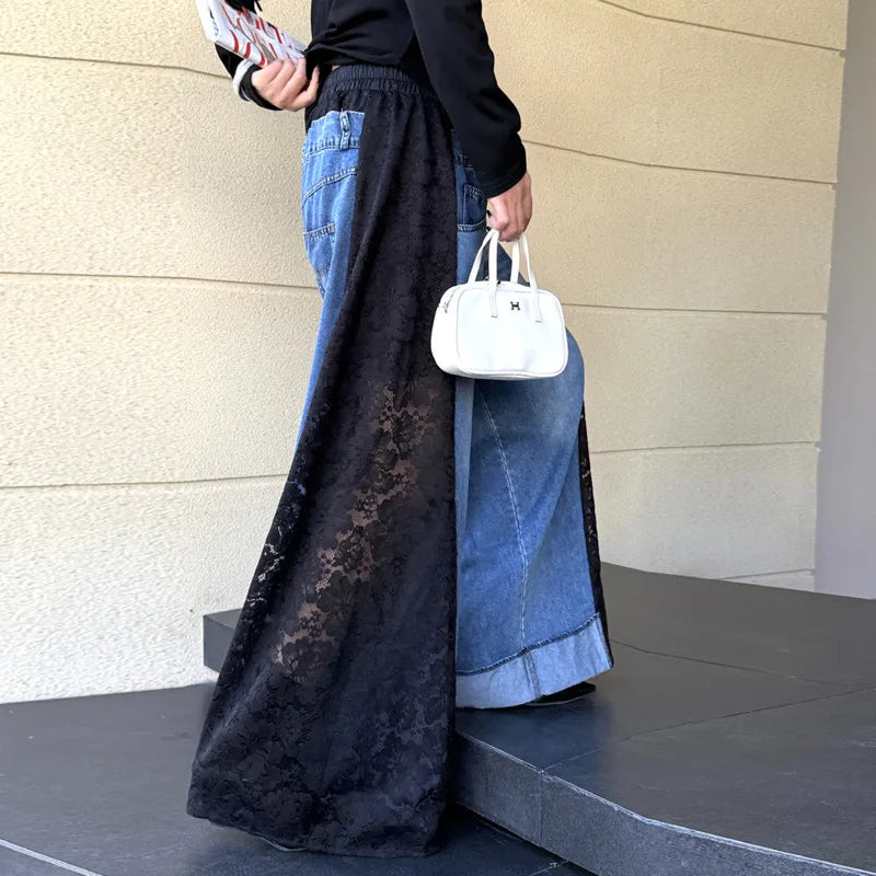 A person wearing a long black lace overlay skirt over blue wide-leg denim jeans, holding a small white handbag, standing on dark steps against a beige textured wall.
