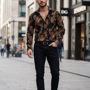A man wearing a long-sleeve button-up shirt with a black base and an intricate paisley pattern in red, gold, and beige, paired with black pants and accessories.