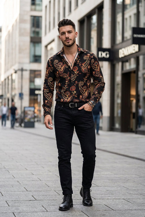 A man wearing a long-sleeve button-up shirt with a black base and an intricate paisley pattern in red, gold, and beige, paired with black pants and accessories.