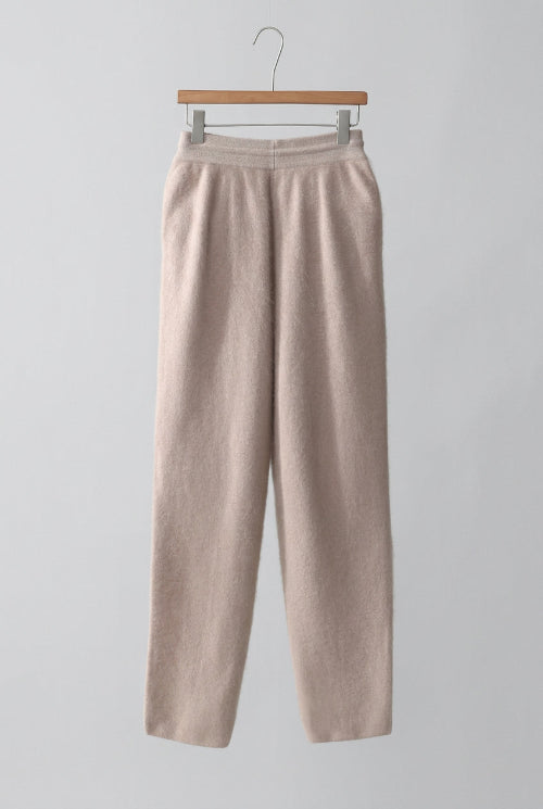 A pair of soft, light beige knit pants hanging on a wooden hanger inside a wooden wardrobe or closet with white drawers.