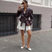 Woman wearing a glossy dark brown belted jacket with wide lapels over a white dress, holding a matching clutch on a sidewalk