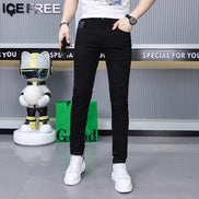 A person modeling slim-fit black jeans with a white t-shirt and white sneakers, standing in front of a gray wall with promotional text and a small robot toy.