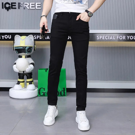 A person modeling slim-fit black jeans with a white t-shirt and white sneakers, standing in front of a gray wall with promotional text and a small robot toy.