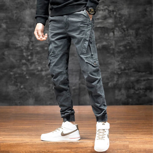 Front view of a model wearing gray cargo pants with angled thigh pockets and gathered ankle cuffs, paired with white high-top sneakers against a dark wall.