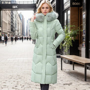 A woman wearing a long, light sage green puffer coat with a large faux-fur trimmed collar stands on a city street. 