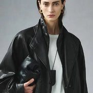 Woman wearing a black faux leather jacket open over a white top, showing layered collars and holding a black handbag against a gray background