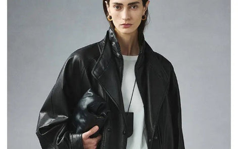 Woman wearing a black faux leather jacket open over a white top, showing layered collars and holding a black handbag against a gray background