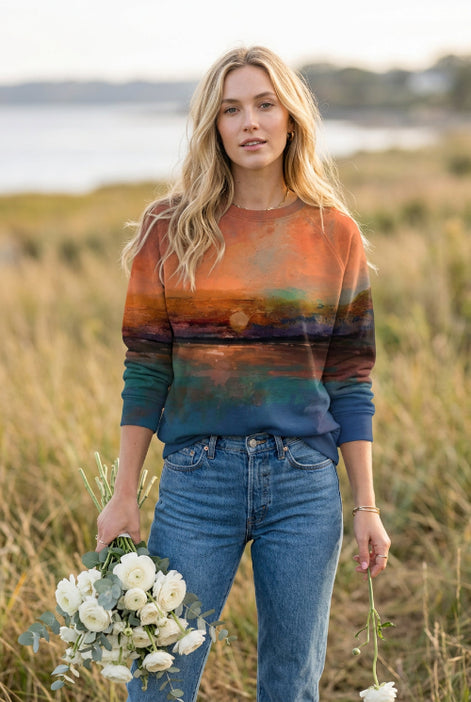 A woman wearing a long-sleeve crewneck sweatshirt with a scenic sunset landscape print, paired with blue jeans and holding a small bouquet of white roses.