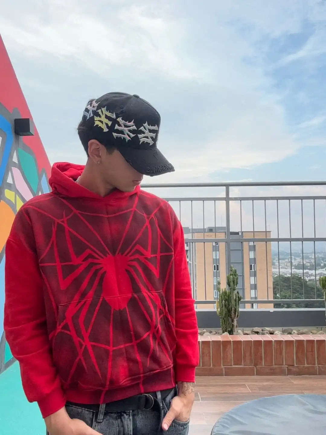 Person wearing a red hoodie with spider web design and black cap on a rooftop with cityscape background