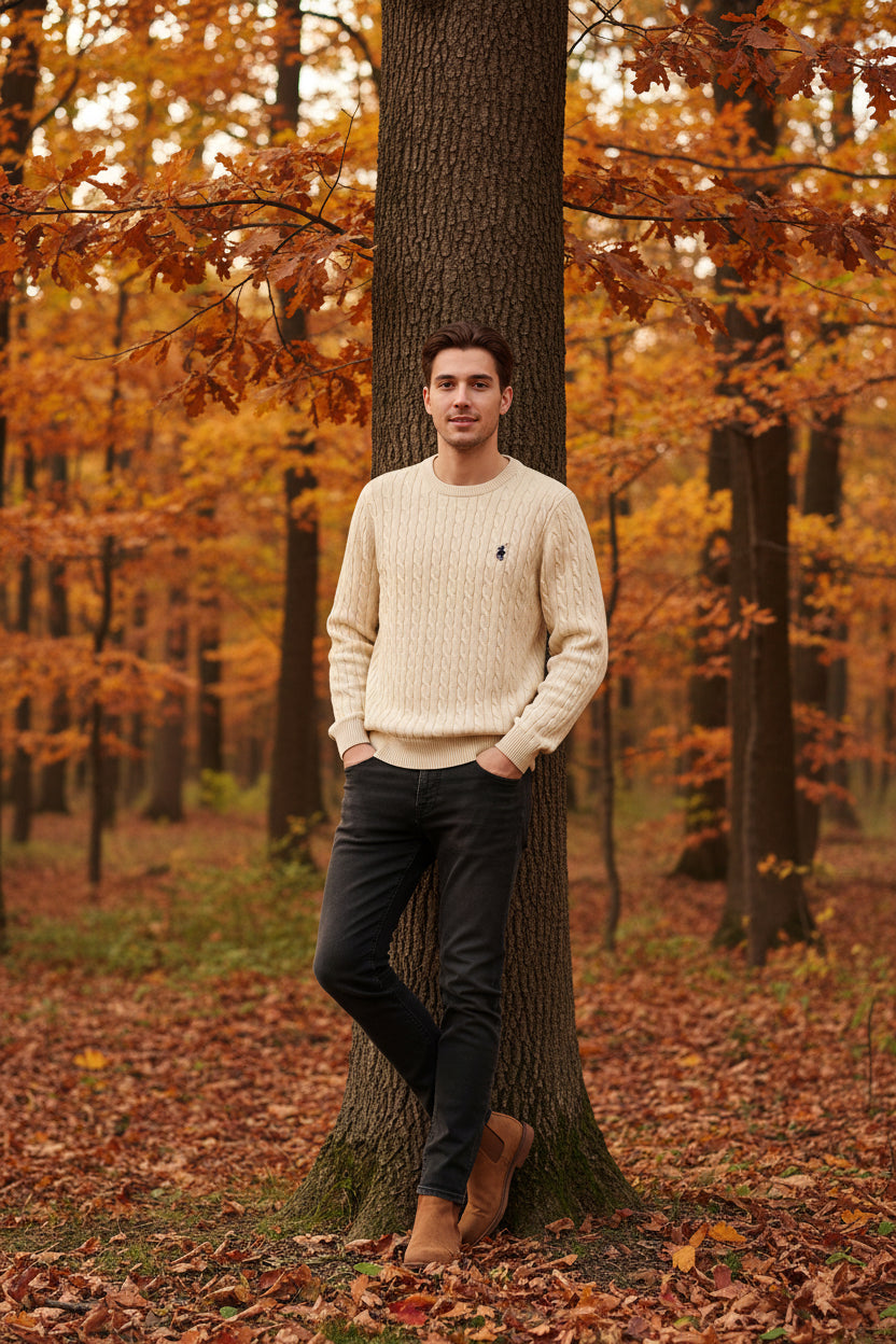 Beige cable-knit sweater with a brand logo held by a hand, on a wooden background.
