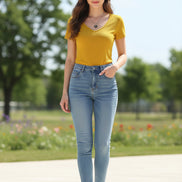A bright yellow short-sleeve V-neck knit top hanging on a white hanger, with a hand holding the hem and dark jeans partially visible below. A necklace with a black heart pendant is draped over the neckline.