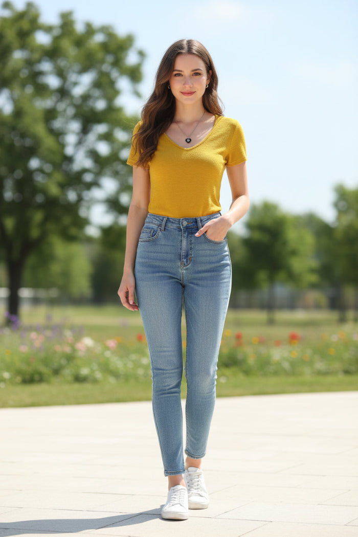 A bright yellow short-sleeve V-neck knit top hanging on a white hanger, with a hand holding the hem and dark jeans partially visible below. A necklace with a black heart pendant is draped over the neckline.