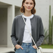 A woman wearing a gray knit button-front cardigan sweater, holding a white phone to take a mirror selfie in a modern indoor setting.