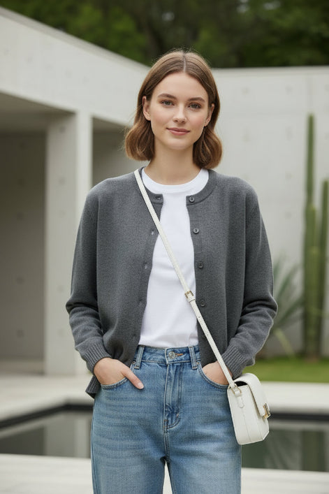 A woman wearing a gray knit button-front cardigan sweater, holding a white phone to take a mirror selfie in a modern indoor setting.