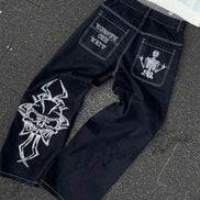 Back view of black wide-leg jeans laid flat, featuring white lettering and a skeleton graphic on the back pockets, white contrast stitching, and a large white cross-style design on one leg.