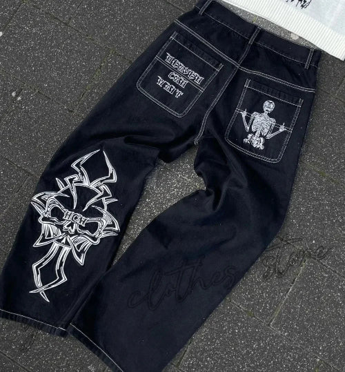 Back view of black wide-leg jeans laid flat, featuring white lettering and a skeleton graphic on the back pockets, white contrast stitching, and a large white cross-style design on one leg.