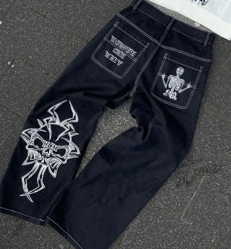 Back view of black wide-leg jeans laid flat, featuring white lettering and a skeleton graphic on the back pockets, white contrast stitching, and a large white cross-style design on one leg.