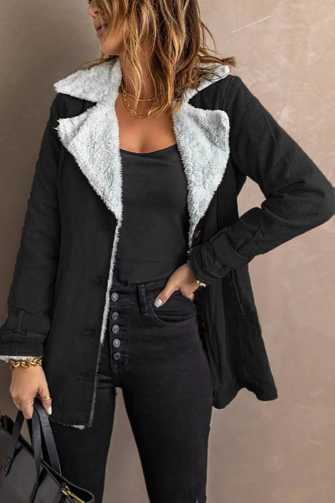Woman wearing a black jacket with white fleece-look lining, wide lapels and buckled cuffs over a black top and jeans
