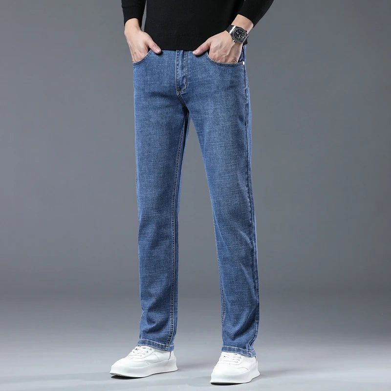 A person wearing blue slim-fit jeans, a black long-sleeve top, and white sneakers, standing against a plain gray studio background with hands in pockets.