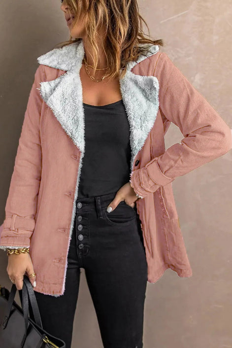 Woman wearing a pink jacket with white fleece-look lining, wide lapels and buckled cuffs over a black top and jeans