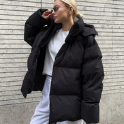 Person wearing a black puffer jacket and white pants against a gray wall.