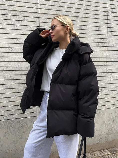 Person wearing a black puffer jacket and white pants against a gray wall.