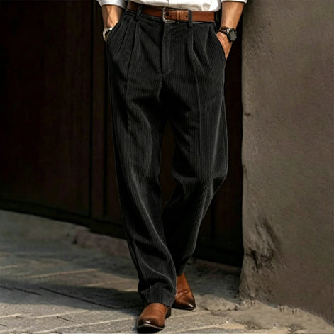 Person wearing dark pleated pants and brown shoes against a textured wall.