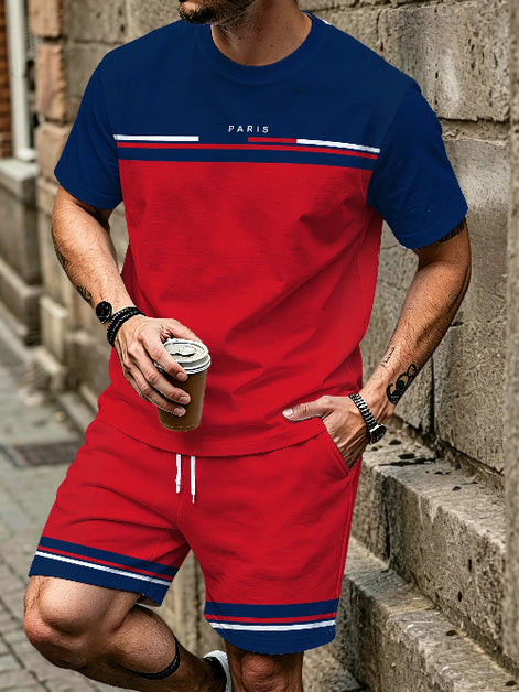 A man wearing a matching red and navy blue athletic set with white stripes and 'PARIS' text on the chest, standing against a stone wall and holding a coffee cup.