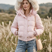 A light pink puffer jacket with a fluffy cream faux-fur trimmed hood, laid flat on top of blue jeans against a white lace-patterned fabric background.