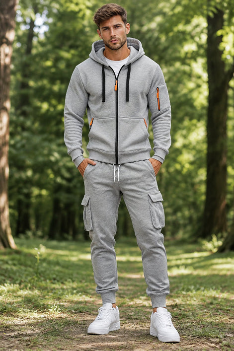 A man wearing a light gray zip-up hoodie and matching gray cargo sweatpants stands in a forest setting. 