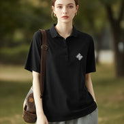A woman wearing a black short-sleeve polo shirt with a small white geometric logo on the left chest, paired with light gray bottoms and carrying a brown shoulder bag.