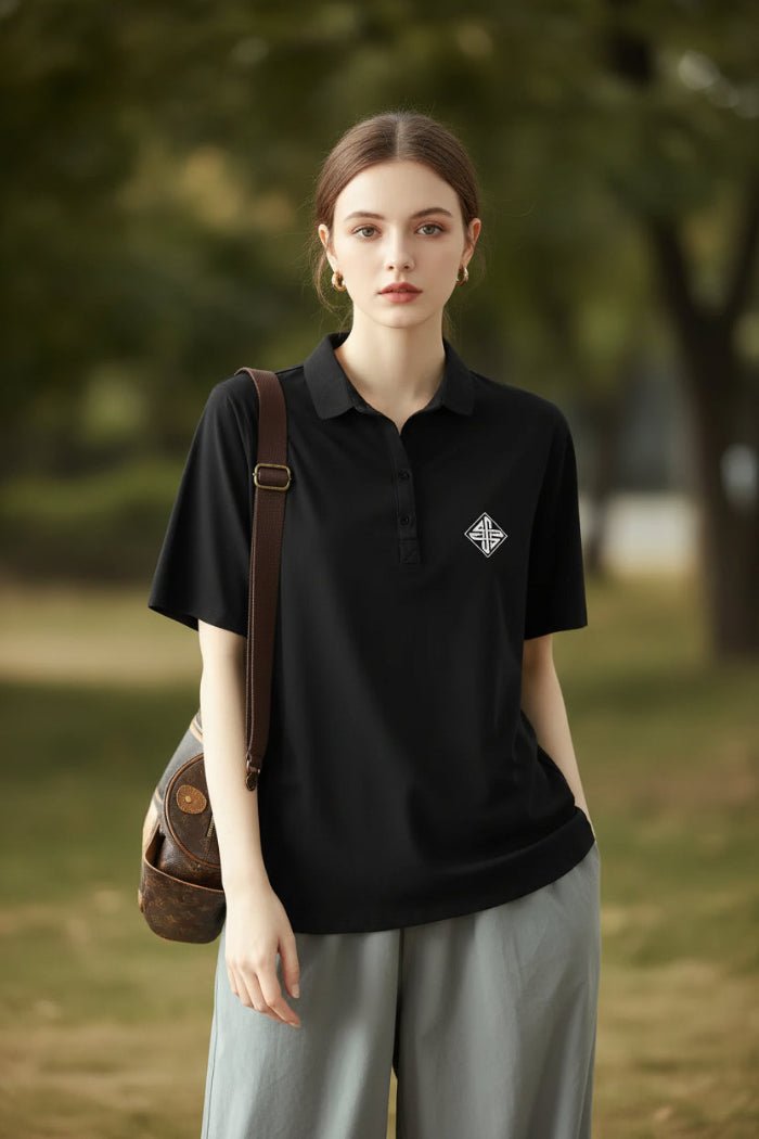 A woman wearing a black short-sleeve polo shirt with a small white geometric logo on the left chest, paired with light gray bottoms and carrying a brown shoulder bag.