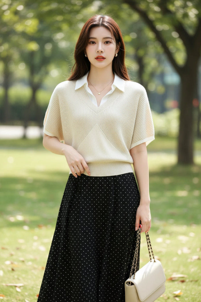 A woman wearing a cream-colored short-sleeve knit polo sweater with a white collar, paired with a black polka-dot skirt and carrying a black shoulder bag.