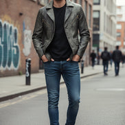 A men's dark gray faux-leather blazer jacket with a textured finish, featuring notched lapels, a three-button front closure, and diagonal zippered chest pockets, displayed on a black shirt against a tan background.