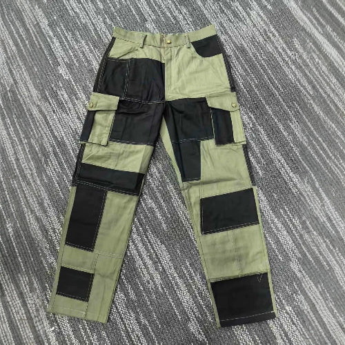 Front view of olive green and black color-block cargo pants laid flat on a gray patterned surface, showing belt loops, a button waistband, and flap thigh pockets.